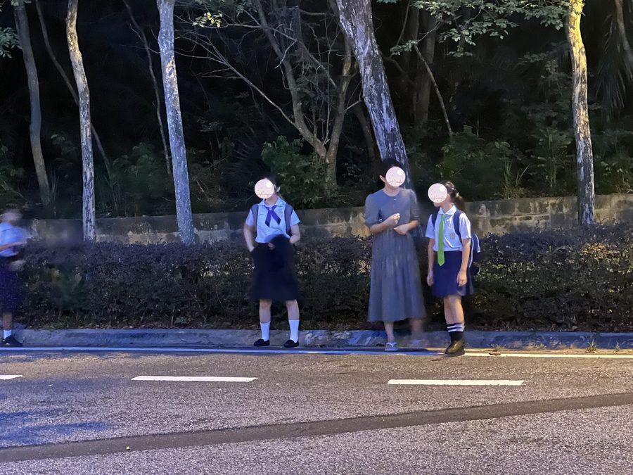 Students in uniform, standing on a road surrounded by green.