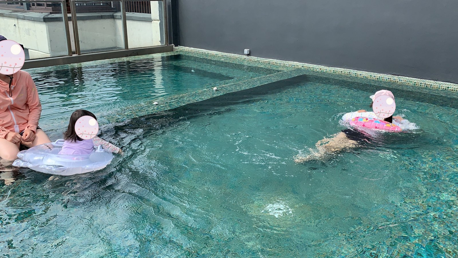 プールサイドで浮き輪の子どもに手を差し伸べる様子。奥では別の子どもも泳いでいる